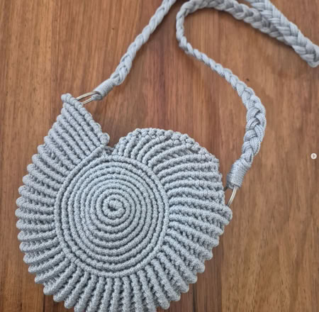 A round, spiral-patterned crocheted bag with a braided strap, placed on a wooden surface.