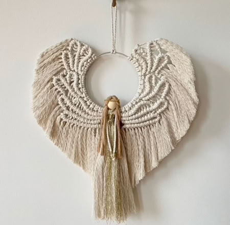 A macramé wall hanging shaped like an angel with a circular center, intricate patterns, and long tassels.
