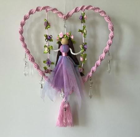 A heart-shaped wall hanging with a braided pink border, featuring a doll in a purple dress with long hair and a flower crown. The doll is surrounded by small flowers and crystal-like ornaments.